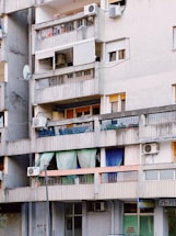 An urban apartment building with several balconies featuring a variety of items such as colorful curtains, potted plants, air conditioning units, and satellite dishes. The structure appears aged, with gray concrete walls and signs of wear.