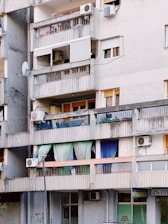An urban apartment building with several balconies featuring a variety of items such as colorful curtains, potted plants, air conditioning units, and satellite dishes. The structure appears aged, with gray concrete walls and signs of wear.