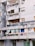 An urban apartment building with several balconies featuring a variety of items such as colorful curtains, potted plants, air conditioning units, and satellite dishes. The structure appears aged, with gray concrete walls and signs of wear.