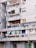 An urban apartment building with several balconies featuring a variety of items such as colorful curtains, potted plants, air conditioning units, and satellite dishes. The structure appears aged, with gray concrete walls and signs of wear.