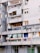 An urban apartment building with several balconies featuring a variety of items such as colorful curtains, potted plants, air conditioning units, and satellite dishes. The structure appears aged, with gray concrete walls and signs of wear.
