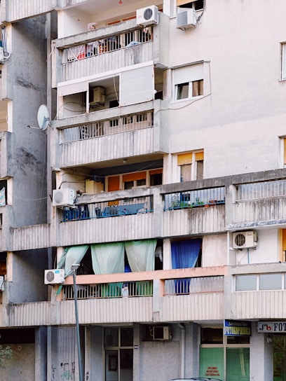 An urban apartment building with several balconies featuring a variety of items such as colorful curtains, potted plants, air conditioning units, and satellite dishes. The structure appears aged, with gray concrete walls and signs of wear.