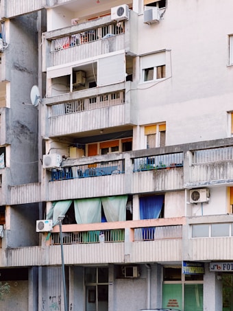 An urban apartment building with several balconies featuring a variety of items such as colorful curtains, potted plants, air conditioning units, and satellite dishes. The structure appears aged, with gray concrete walls and signs of wear.