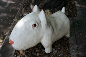 A concrete sculpture of a dog with a large body, short legs, and prominent facial features, placed on a bed of small rocks. The sculpture is white with a red nose and black eyes, situated in an outdoor environment with some foliage and a tree trunk nearby.