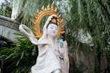 An exquisite statue of a figure with a serene expression, adorned with a golden halo and ornate clothing. The statue is surrounded by lush green foliage, adding to the peaceful and spiritual atmosphere. Intricate details like the flowing garments and delicate facial features enhance its beauty.
