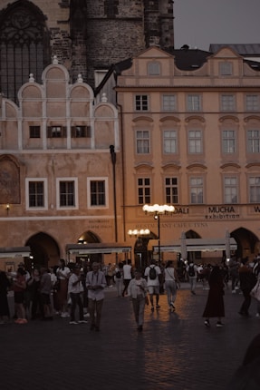 A lively scene in a historic European square with beautiful, warm-toned buildings and ornate architectural details. Many people are walking and gathering, enjoying the square in the evening light. Street lamps illuminate the area with a soft glow, adding a cozy atmosphere.