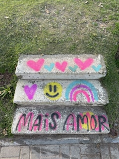 Concrete steps adorned with vibrant graffiti, featuring colorful hearts, a yellow smiley face, and a rainbow. Below these, the words 'MAIS AMOR' are spray-painted in bold, bright colors. The steps are surrounded by green grass on either side.