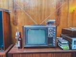 A vintage television with a wood panel exterior sits on a wooden cabinet. The TV has a small screen with dials on the side and two antennae extending upwards. On top of the television are several VHS tapes. To the left of the television, there are open and unopened glass soda bottles. On the right, more VHS tapes are stacked beside a stereo system. The setting features wooden paneled walls, evoking a retro atmosphere.