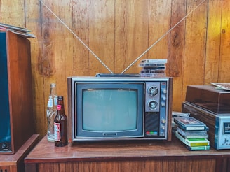 A vintage television with a wood panel exterior sits on a wooden cabinet. The TV has a small screen with dials on the side and two antennae extending upwards. On top of the television are several VHS tapes. To the left of the television, there are open and unopened glass soda bottles. On the right, more VHS tapes are stacked beside a stereo system. The setting features wooden paneled walls, evoking a retro atmosphere.