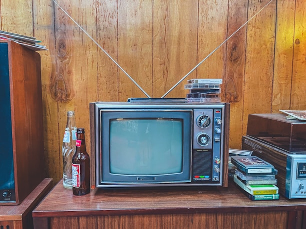 A vintage television with a wood panel exterior sits on a wooden cabinet. The TV has a small screen with dials on the side and two antennae extending upwards. On top of the television are several VHS tapes. To the left of the television, there are open and unopened glass soda bottles. On the right, more VHS tapes are stacked beside a stereo system. The setting features wooden paneled walls, evoking a retro atmosphere.