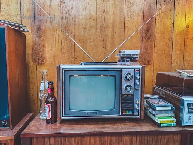 A vintage television with a wood panel exterior sits on a wooden cabinet. The TV has a small screen with dials on the side and two antennae extending upwards. On top of the television are several VHS tapes. To the left of the television, there are open and unopened glass soda bottles. On the right, more VHS tapes are stacked beside a stereo system. The setting features wooden paneled walls, evoking a retro atmosphere.