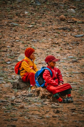 Two children are sitting on a rocky, barren landscape. They are wearing brightly colored clothes, with one in a yellow outfit and red hat, and the other in a red outfit with a cap. Each child has a backpack, suggesting they might be on a hike or an outdoor trip.