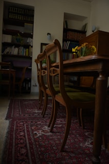 A room with wooden chairs arranged alongside an ornate, patterned red carpet. Bookshelves filled with books and files line the walls, while a vase with yellow flowers sits on the table, adding a touch of color to the subdued surroundings.