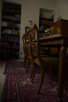 A room with wooden chairs arranged alongside an ornate, patterned red carpet. Bookshelves filled with books and files line the walls, while a vase with yellow flowers sits on the table, adding a touch of color to the subdued surroundings.