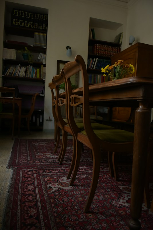 A room with wooden chairs arranged alongside an ornate, patterned red carpet. Bookshelves filled with books and files line the walls, while a vase with yellow flowers sits on the table, adding a touch of color to the subdued surroundings.