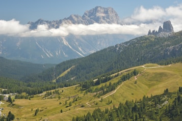 A picturesque mountain landscape featuring a range of green hills, dense forests, and a backdrop of towering rocky peaks partially obscured by a layer of clouds. Winding trails can be seen cutting through the verdant terrain, inviting exploration.