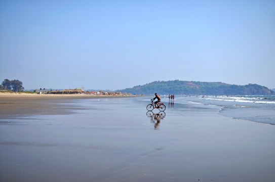 A serene beach scene with a person riding a bicycle along the smooth, reflective shoreline. In the background, people are walking and enjoying the beach. The coastline curves gently with distant hills and a clear blue sky, creating a peaceful atmosphere.