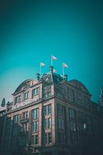 A large, historic building with intricate architectural details, featuring a sign that reads DE BIJENKORF. The structure has a sloped roof adorned with small spires and several flags waving at the top. The sky is clear with a teal hue, and the building is partially shadowed, likely due to the time of day.