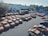 A large number of orange taxis are parked in an open lot. The lot is divided into several rows with taxis neatly arranged. There are a few structures in the background, including a few small buildings and a yellow container. Trees and industrial buildings are visible in the background.