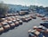A large number of orange taxis are parked in an open lot. The lot is divided into several rows with taxis neatly arranged. There are a few structures in the background, including a few small buildings and a yellow container. Trees and industrial buildings are visible in the background.