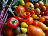 A variety of fresh vegetables, including different types of tomatoes, red chili peppers, and bell peppers. The fruits and vegetables are displayed with vibrant colors and appear ripe and fresh. Green leaves can be seen in the background, adding to the lush appearance.