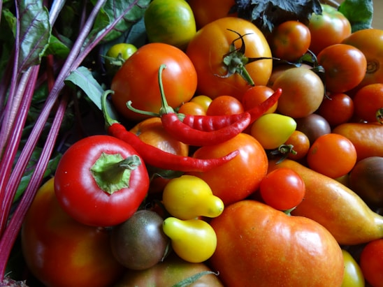 A variety of fresh vegetables, including different types of tomatoes, red chili peppers, and bell peppers. The fruits and vegetables are displayed with vibrant colors and appear ripe and fresh. Green leaves can be seen in the background, adding to the lush appearance.