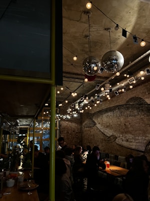 A dimly-lit bar or caf&eacute; with exposed brick walls and industrial decor. Hanging lights and disco balls create a cozy and inviting atmosphere. The space is filled with people seated at tables and the bar, engaged in conversation. A large mural of a fish is painted on one of the walls.