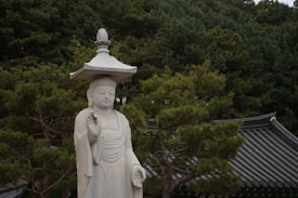 A stone statue of Buddha stands peacefully among lush green pine trees with a traditional temple rooftop in the background. The Buddha is depicted with a serene expression and holds a unique umbrella-like structure above its head.