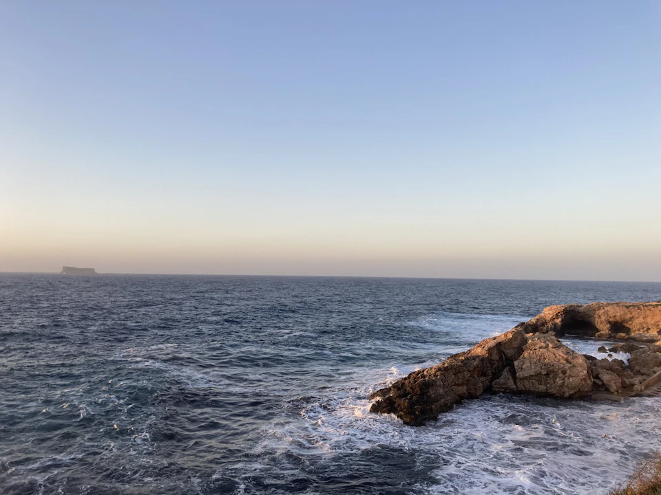 Rocky cliff overlooks serene ocean with calm waters and distant ships in blue sky