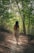 A smiling woman walking barefoot on a sunlit forest path wearing minimalist macakisses shoes.