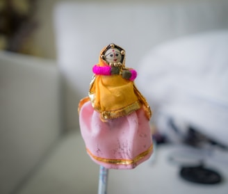 A small, handcrafted doll wearing traditional attire, including a bright orange dress with gold accents and a pink lower portion. The doll has a painted face with intricate detailing and is adorned with beads and a small pink sash.