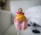 A small, handcrafted doll wearing traditional attire, including a bright orange dress with gold accents and a pink lower portion. The doll has a painted face with intricate detailing and is adorned with beads and a small pink sash.