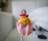 A small, handcrafted doll wearing traditional attire, including a bright orange dress with gold accents and a pink lower portion. The doll has a painted face with intricate detailing and is adorned with beads and a small pink sash.