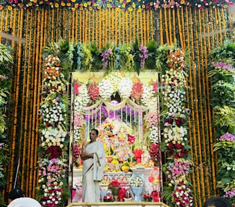 The image depicts a vibrant and elaborately decorated altar filled with colorful flowers arranged in various patterns. A person in traditional attire stands in front, surrounded by floral garlands. The altar centerpiece consists of intricately dressed figures, possibly deities, with offerings placed in front.
