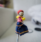 A small handmade doll stands prominently, featuring a fabric face with drawn-on facial features. It is dressed in colorful traditional clothing, including a red headpiece, a pink polka dot upper garment, and a blue skirt with orange accents. The doll seems to be crafted from various materials including fabric and yarn.