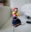 A small handmade doll stands prominently, featuring a fabric face with drawn-on facial features. It is dressed in colorful traditional clothing, including a red headpiece, a pink polka dot upper garment, and a blue skirt with orange accents. The doll seems to be crafted from various materials including fabric and yarn.