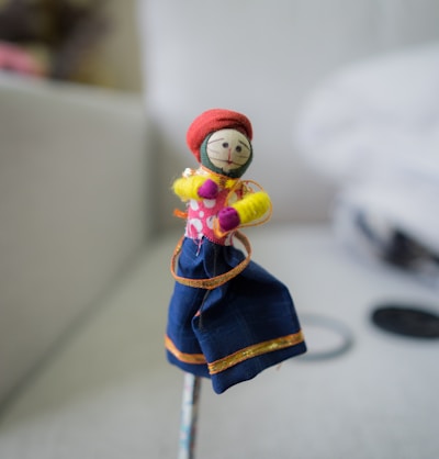 A small handmade doll stands prominently, featuring a fabric face with drawn-on facial features. It is dressed in colorful traditional clothing, including a red headpiece, a pink polka dot upper garment, and a blue skirt with orange accents. The doll seems to be crafted from various materials including fabric and yarn.