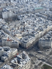 Aerial view of a densely populated urban area featuring a grid-like pattern of buildings. The architecture is characterized by uniformity in design with grey rooftops and a few streets visible. Sparse trees line the boulevards and there are minimal vehicles and people scattered throughout the scene.