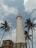 A tall, white cylindrical lighthouse stands majestically against a partly cloudy sky. It is flanked by several tall palm trees swaying gently in the breeze. Adjacent to the lighthouse, there is a small building with a ladder leading up to the roof.
