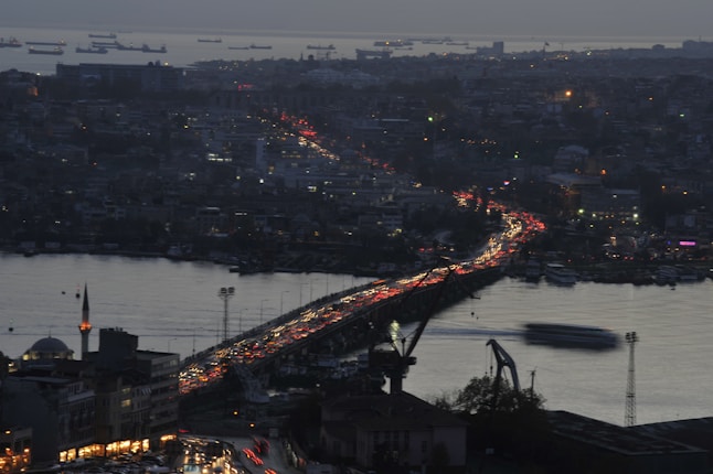 A bustling cityscape at dusk with a bridge full of traffic spanning across a body of water. The city lights create a vibrant and energetic atmosphere, while numerous ships are visible in the distance on the water. Buildings line the waterfront, and a mosque with a prominent minaret stands in the foreground.