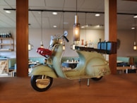 A vintage-style model scooter displayed on a wooden shelf in a cozy dining area. The scooter is painted in pastel green, with red coiled wire attached to the front. It has a classic design with a small luggage rack on the back. The background consists of a softly lit interior with pendant lights and wooden furniture.