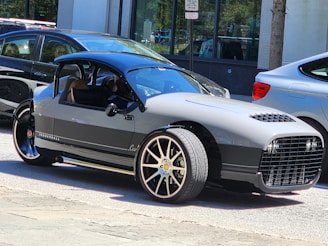 A sleek three-wheeled vehicle with a modern, open-top design is parked on the street. It features a streamlined body, prominent front grille, and distinct alloy wheels, surrounded by other parked cars and urban buildings.
