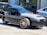 A sleek three-wheeled vehicle with a modern, open-top design is parked on the street. It features a streamlined body, prominent front grille, and distinct alloy wheels, surrounded by other parked cars and urban buildings.