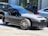 A sleek three-wheeled vehicle with a modern, open-top design is parked on the street. It features a streamlined body, prominent front grille, and distinct alloy wheels, surrounded by other parked cars and urban buildings.