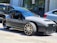 A sleek three-wheeled vehicle with a modern, open-top design is parked on the street. It features a streamlined body, prominent front grille, and distinct alloy wheels, surrounded by other parked cars and urban buildings.
