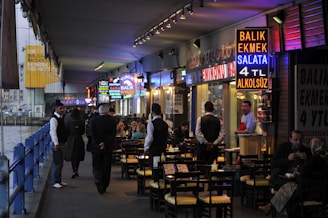 A bustling waterfront restaurant area underneath a bridge, with vibrant neon signs advertising food such as fish sandwiches. Several people are seated at outdoor tables enjoying their meals, while waitstaff in uniforms attend to them. The setting appears lively with a mix of locals and tourists.
