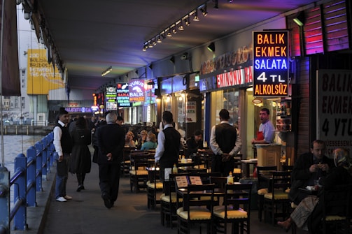 A bustling waterfront restaurant area underneath a bridge, with vibrant neon signs advertising food such as fish sandwiches. Several people are seated at outdoor tables enjoying their meals, while waitstaff in uniforms attend to them. The setting appears lively with a mix of locals and tourists.