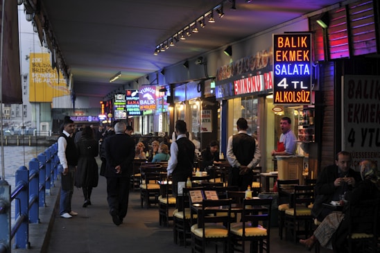A bustling waterfront restaurant area underneath a bridge, with vibrant neon signs advertising food such as fish sandwiches. Several people are seated at outdoor tables enjoying their meals, while waitstaff in uniforms attend to them. The setting appears lively with a mix of locals and tourists.