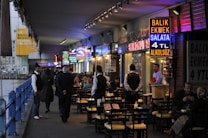 A bustling waterfront restaurant area underneath a bridge, with vibrant neon signs advertising food such as fish sandwiches. Several people are seated at outdoor tables enjoying their meals, while waitstaff in uniforms attend to them. The setting appears lively with a mix of locals and tourists.
