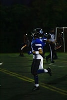 A person in a blue football uniform is running on a field. The player is wearing a helmet with a face guard. Other players and a referee can be seen in the background, all involved in an outdoor sports event during nighttime.